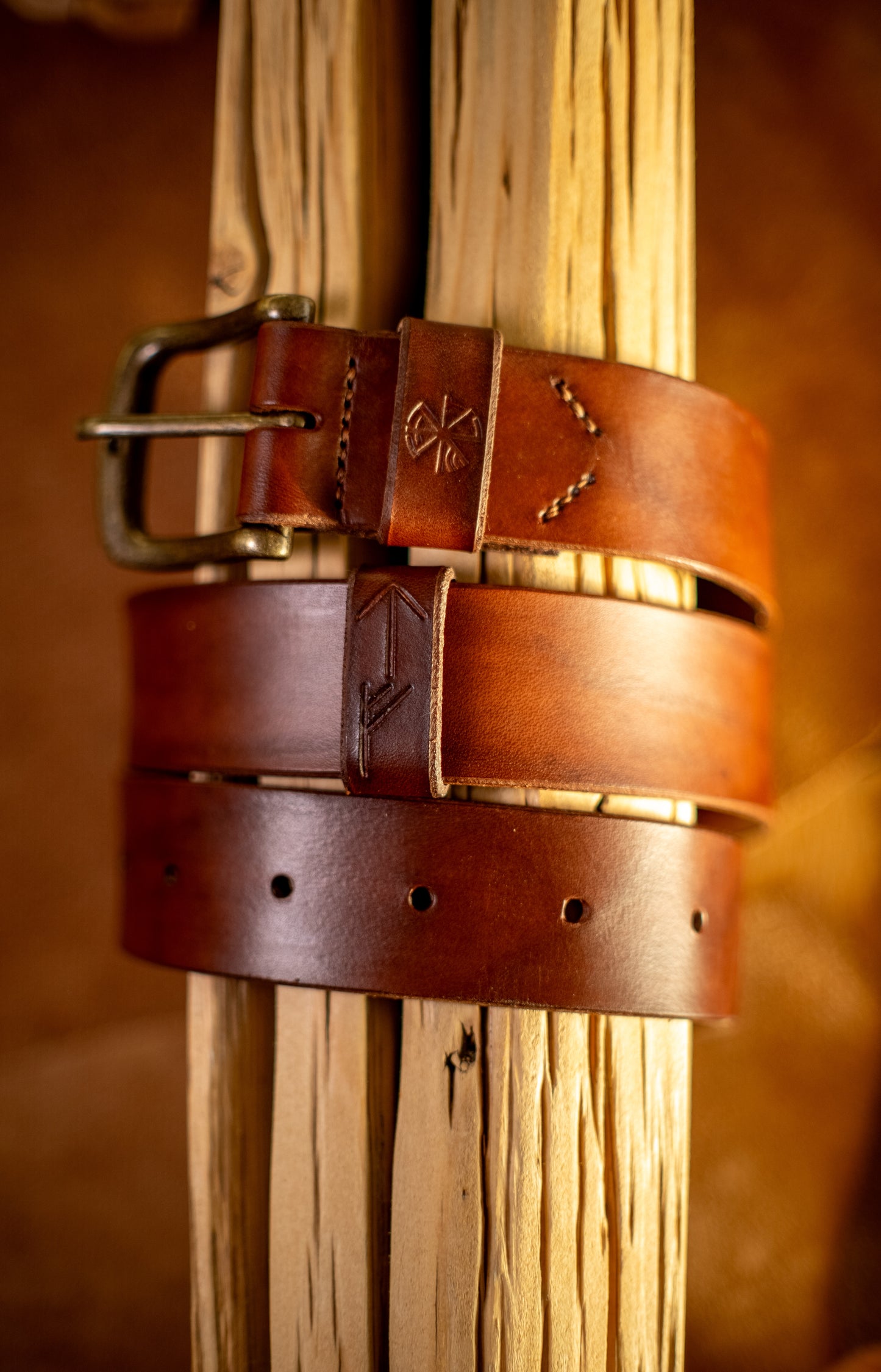 Et håndlaget lærbelte, Gleipnir 40mm, i en dyp mørk brun farge, elegant viklet rundt rustikt treverk. Beltet har en solid messingspenne, håndsydde detaljer og pregede symboler inspirert av norrøn mytologi. Den rike brunfargen og den glatte overflaten understreker beltets høye kvalitet og tidløse design, laget for å vare i generasjoner.