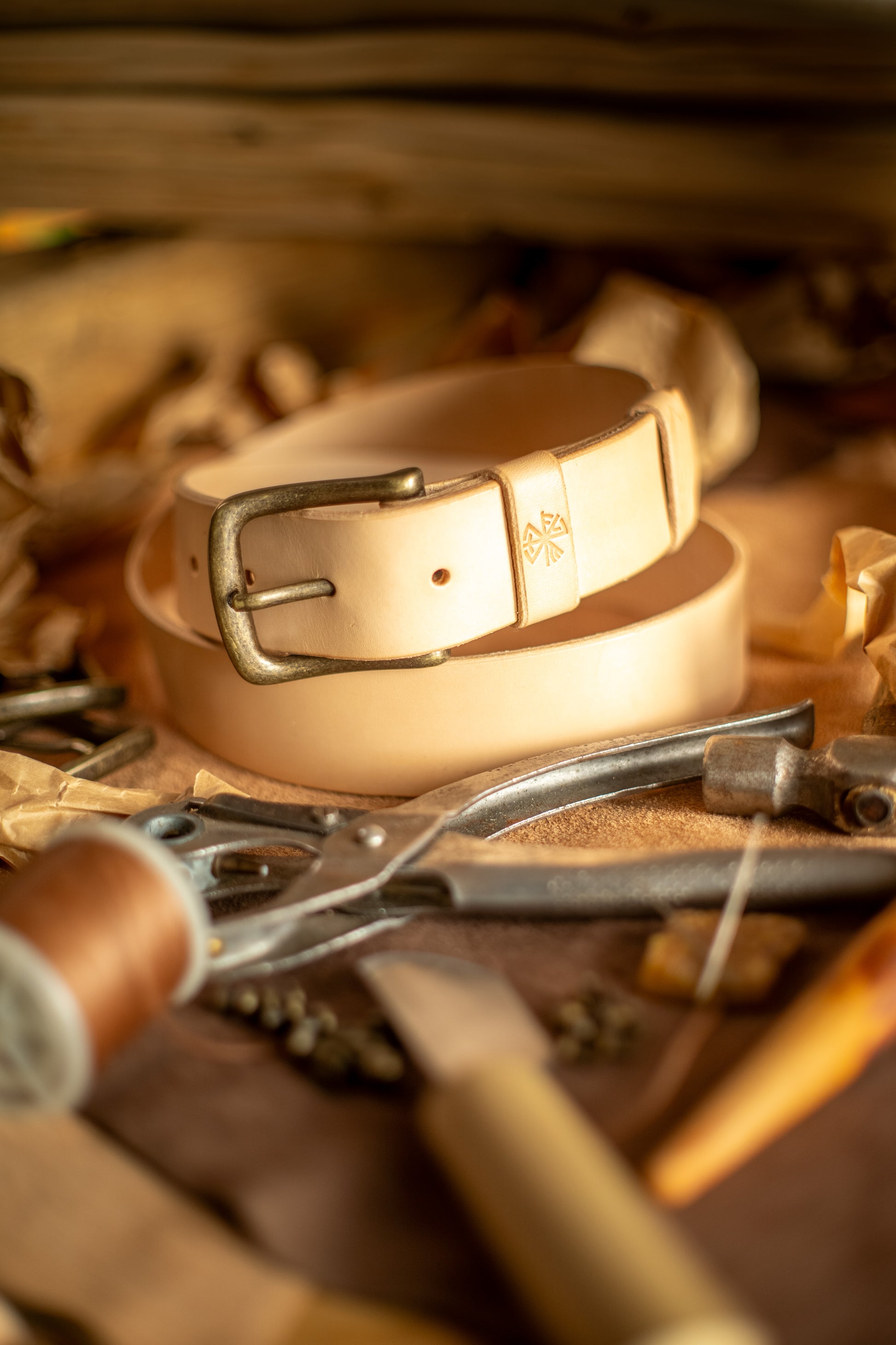 Et håndlaget lærbelte, Gleipnir 40mm, i naturlig ufarget lær med en solid messingspenne. Beltet har et tidløst design med subtile pregede detaljer og er omgitt av tradisjonelle håndverksverktøy. Bildet fremhever den autentiske produksjonsprosessen og kvaliteten på dette slitesterke og bærekraftige tilbehøret, laget for å vare i generasjoner.