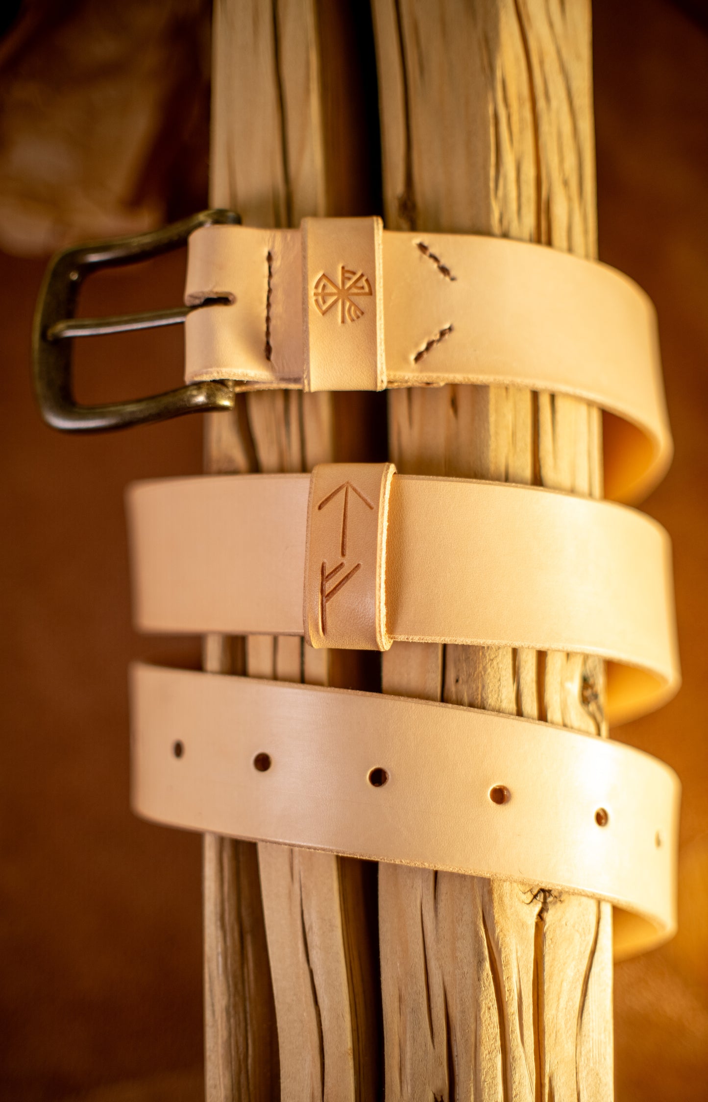 Et håndlaget lærbelte, Gleipnir 40mm, i naturlig ufarget lær, elegant viklet rundt rustikt treverk. Beltet har en solid messingspenne, håndsydde detaljer og pregede symboler inspirert av norrøn mytologi. Den lyse, rå finishen på læret understreker det autentiske håndverket og bærekraftige materialvalget, skapt for å vare i generasjoner.