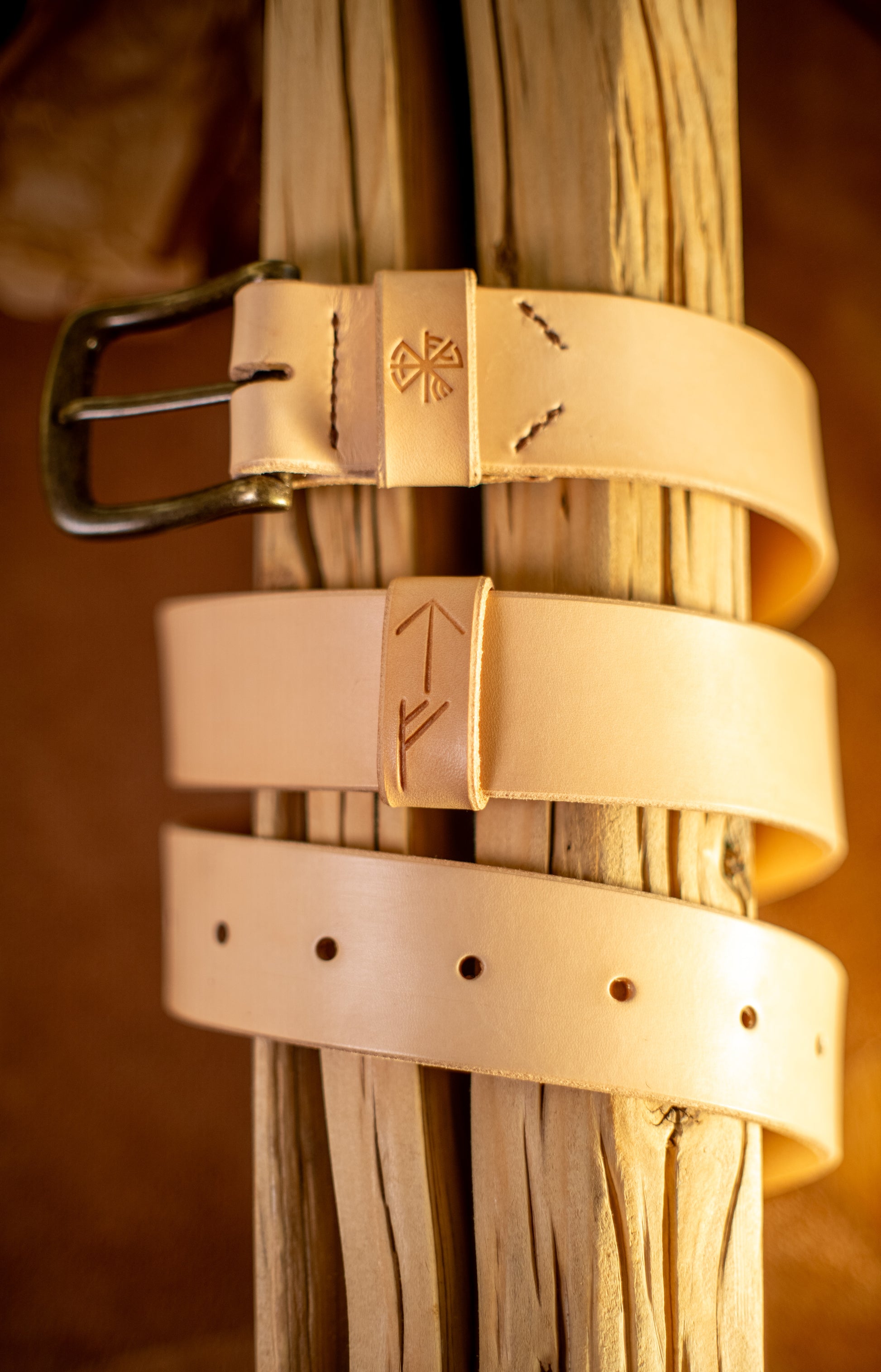 Et håndlaget lærbelte, Gleipnir 40mm, i naturlig ufarget lær, elegant viklet rundt rustikt treverk. Beltet har en solid messingspenne, håndsydde detaljer og pregede symboler inspirert av norrøn mytologi. Den lyse, rå finishen på læret understreker det autentiske håndverket og bærekraftige materialvalget, skapt for å vare i generasjoner.