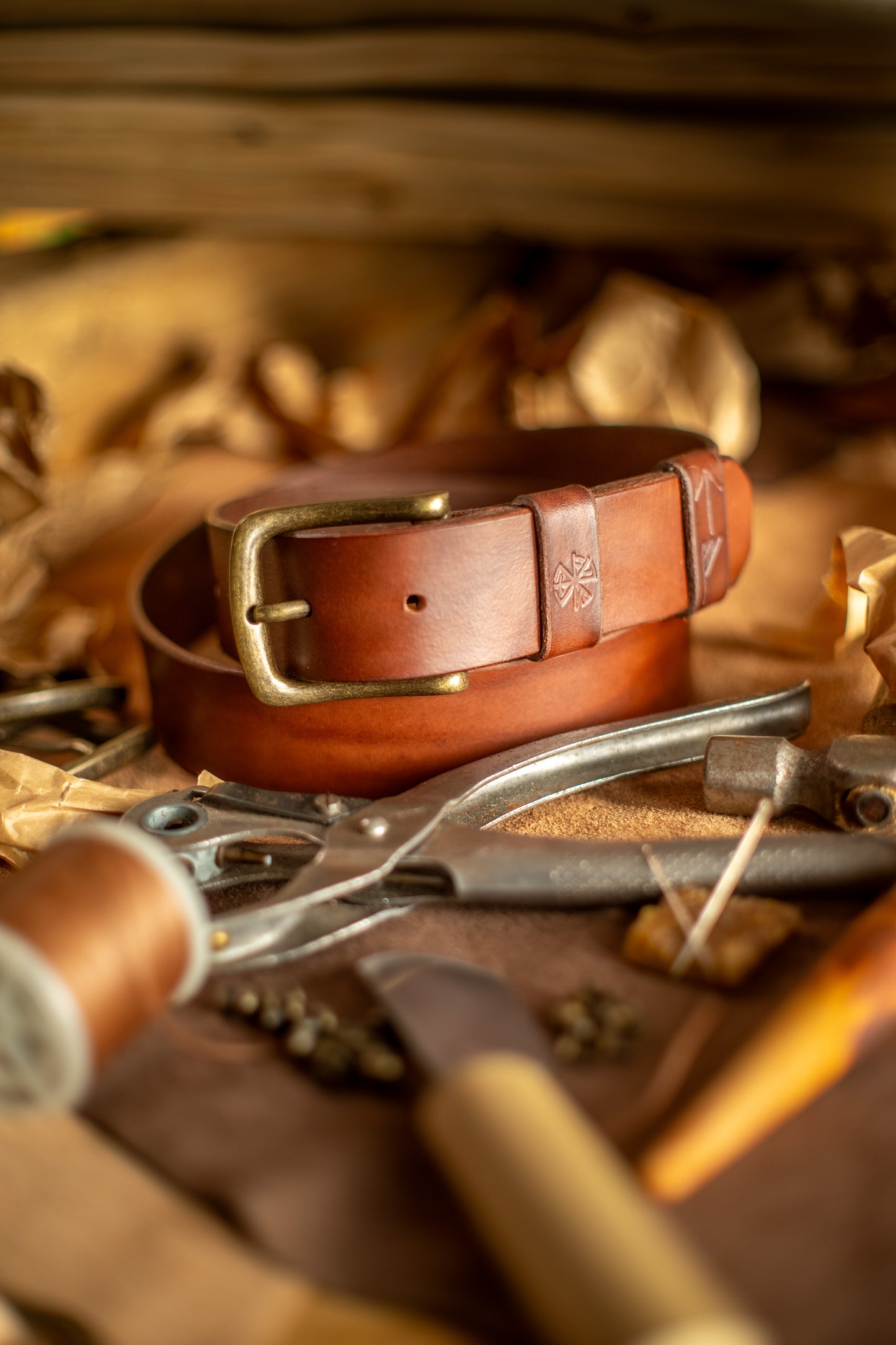Et håndlaget lærbelte, Gleipnir 40mm, i en dyp mørk brun nyanse med en solid messingspenne. Beltet har et tidløst og robust design med pregede detaljer inspirert av norrøn mytologi. Omgitt av håndverksverktøy og materialer, fremhever bildet den autentiske produksjonsprosessen og den høye kvaliteten på dette slitesterke og bærekraftige tilbehøret.
