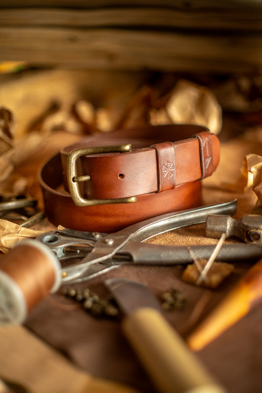 Et håndlaget lærbelte, Gleipnir 40mm, i en dyp mørk brun nyanse med en solid messingspenne. Beltet har et tidløst og robust design med pregede detaljer inspirert av norrøn mytologi. Omgitt av håndverksverktøy og materialer, fremhever bildet den autentiske produksjonsprosessen og den høye kvaliteten på dette slitesterke og bærekraftige tilbehøret.
