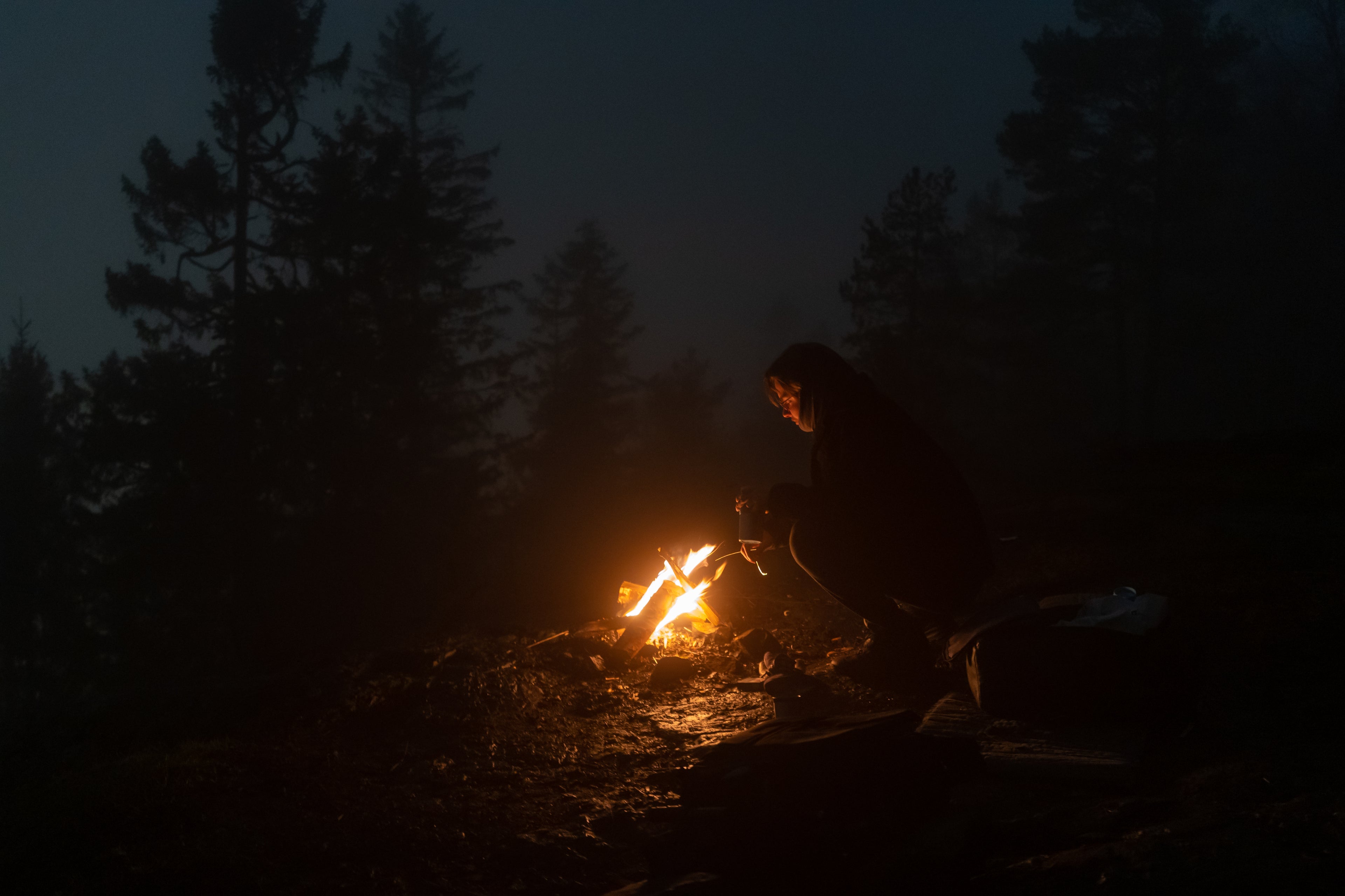 En person sitter ved et leirbål i en mørk skog, omgitt av tåke og høye trær. Flammene kaster et varmt lys som lyser opp ansiktet og omgivelsene, og skaper en stemningsfull kontrast mot den dystre naturen. Bildet fanger essensen av eventyr og friluftsliv, og reflekterer verdiene til Tokt Film og Tokt Supply – autentiske opplevelser, bærekraftige produkter og en tilknytning til naturen.
